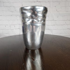 May include: A silver-colored, hammered metal vase with a textured surface. The vase has a wide, irregular top and a tapered body. The vase is displayed on a dark brown wooden table against a white brick wall.