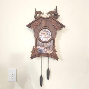 May include: A brown wooden cuckoo clock with a forest scene on the face and two deer figurines on top. The clock has a pendulum with two pinecone weights and is mounted on a cream-colored wall.