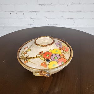 May include: A ceramic bowl with a lid, featuring a floral design in red, yellow, and purple. The bowl has a beige base and a brown rim. The lid has a circular opening. The bowl is on a dark brown surface.
