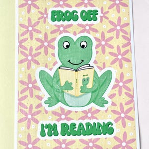 Op de afbeelding: Een wenskaart met een groene kikker die een boek leest, met de tekst "Frog Off I'm Reading". De kikker staat tegen een gele achtergrond met roze bloemmotieven. De kaart heeft een witte rand.