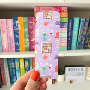 Op de afbeelding: Een rechthoekige boekenlegger met een lavendelkleurige achtergrond en een herhalend patroon van boeken, boekenplanken, koffiekopjes en sterren. De boekenlegger wordt vastgehouden in een hand met oranje nagellak. Boekenplanken vol boeken staan op de achtergrond.