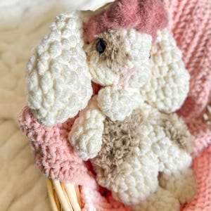 May include: A handmade crochet bunny toy with white, brown, and pink accents. The bunny has long floppy ears and is wearing a pink bow. It rests in a pink crocheted basket, creating a soft and cuddly aesthetic.