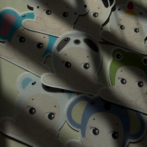 May include: A collection of glittery stickers featuring cartoon characters in animal costumes, including a panda, bear, frog, and elephant. The stickers have a light beige base color with colorful accents.