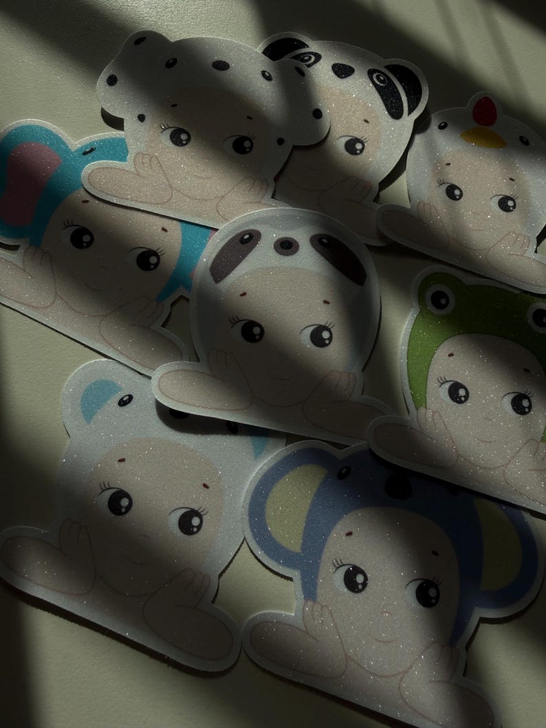 May include: A collection of glittery stickers featuring cartoon characters in animal costumes, including a panda, bear, frog, and elephant. The stickers have a light beige base color with colorful accents.