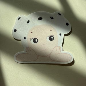 May include: A whimsical sticker featuring a cartoon character with a mushroom cap. The character has a light skin tone, large eyes, and is resting its chin on its hands. The mushroom cap is white with black polka dots.