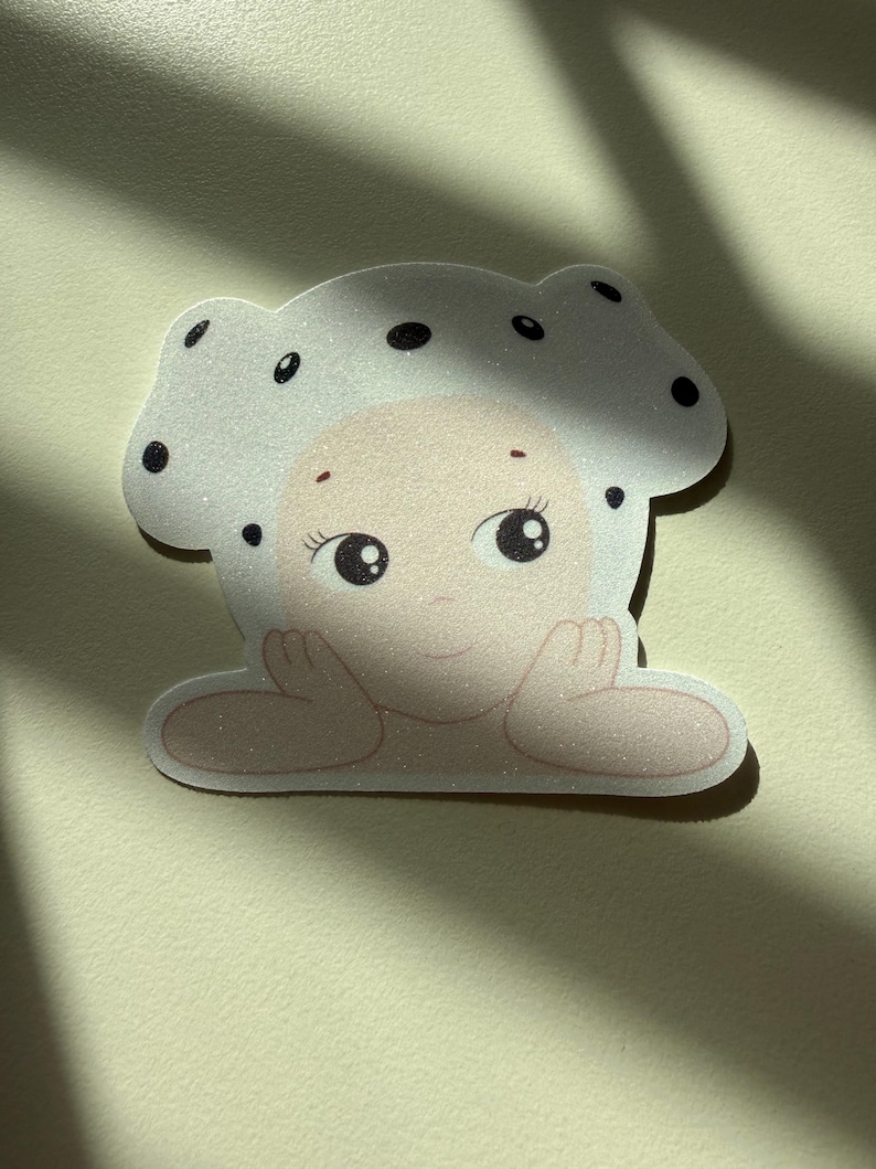 May include: A whimsical sticker featuring a cartoon character with a mushroom cap. The character has a light skin tone, large eyes, and is resting its chin on its hands. The mushroom cap is white with black polka dots.