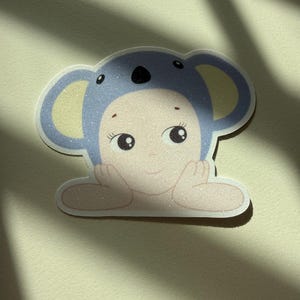 May include: A sticker featuring a baby-faced character in a blue koala hat. The sticker has a glittery finish and a light skin tone. The character's hands are resting on a surface, and it has large, expressive eyes.