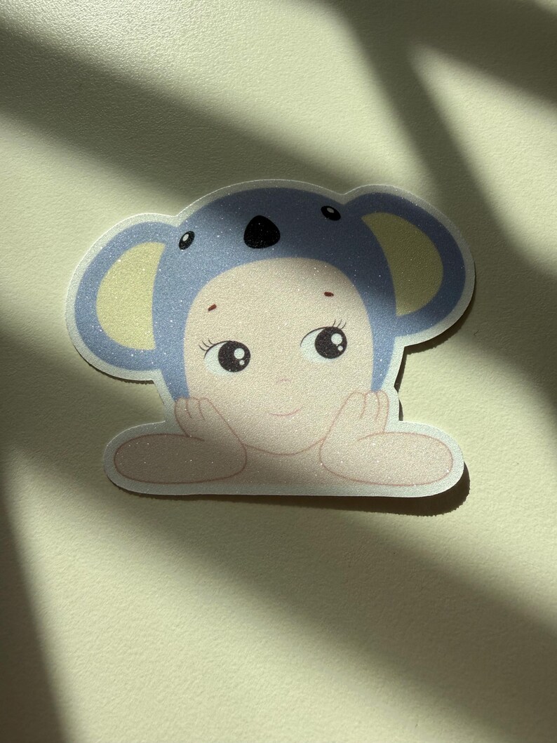 May include: A sticker featuring a baby-faced character in a blue koala hat. The sticker has a glittery finish and a light skin tone. The character's hands are resting on a surface, and it has large, expressive eyes.