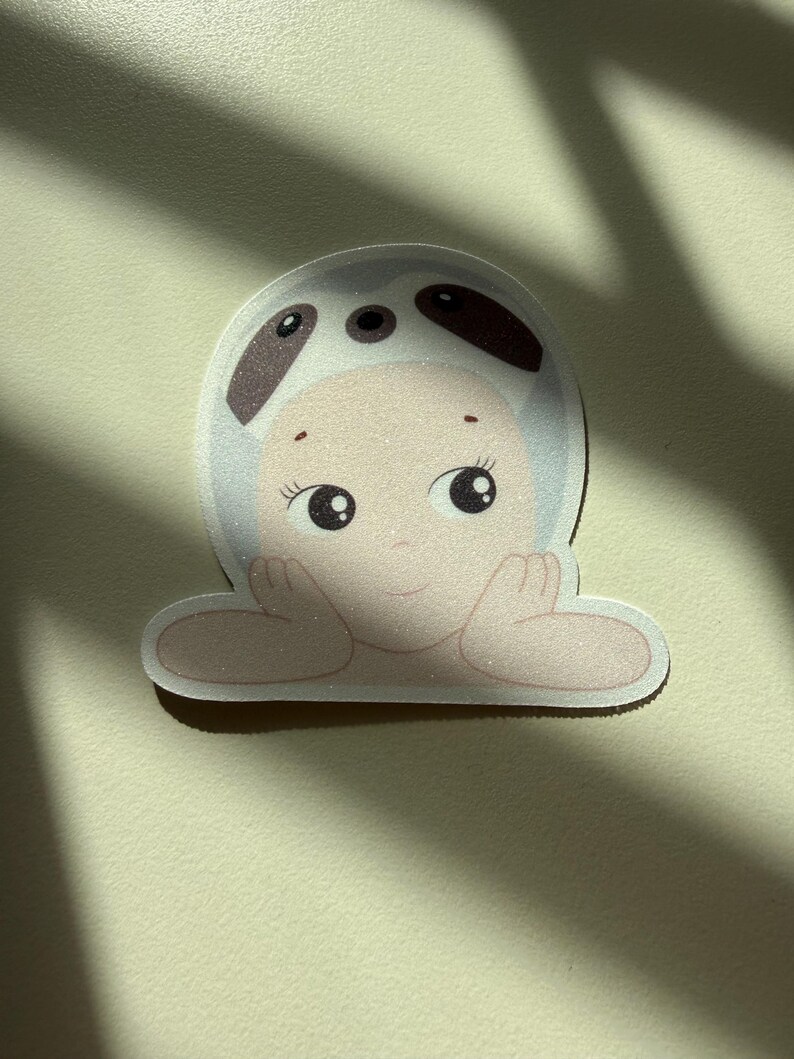 May include: A sticker featuring a cartoon baby with large eyes and a smiling expression. The baby is wearing a white hat with brown spots and is resting its chin on its hands. The sticker has a light pink and beige color scheme.