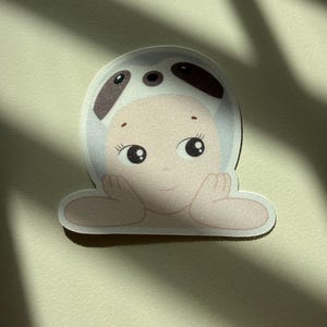 May include: A sticker featuring a cartoon baby wearing a panda hat. The baby has large, expressive eyes and is resting its arms on a surface. The sticker has a white border and a light beige background.