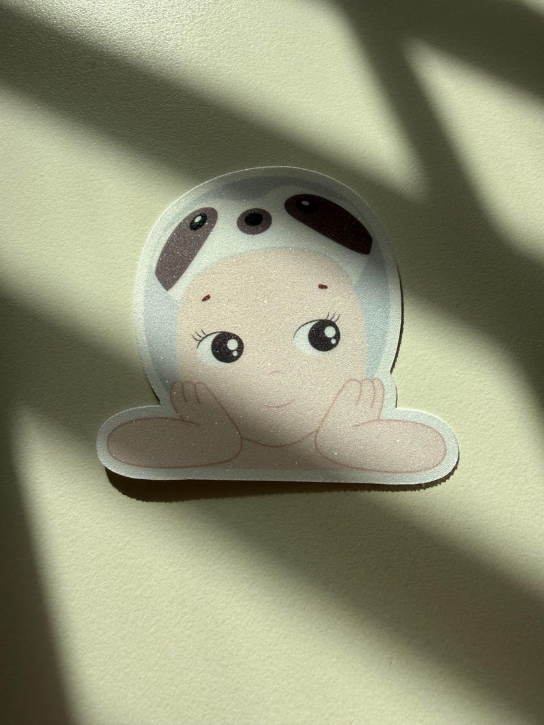 May include: A sticker featuring a cartoon baby wearing a panda hat. The baby has large, expressive eyes and is resting its arms on a surface. The sticker has a white border and a light beige background.