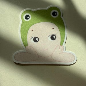 May include: A sticker featuring a cartoon illustration of a person wearing a green frog hat. The person has large, expressive eyes and is resting their chin on their hands. The hat has a glittery texture.