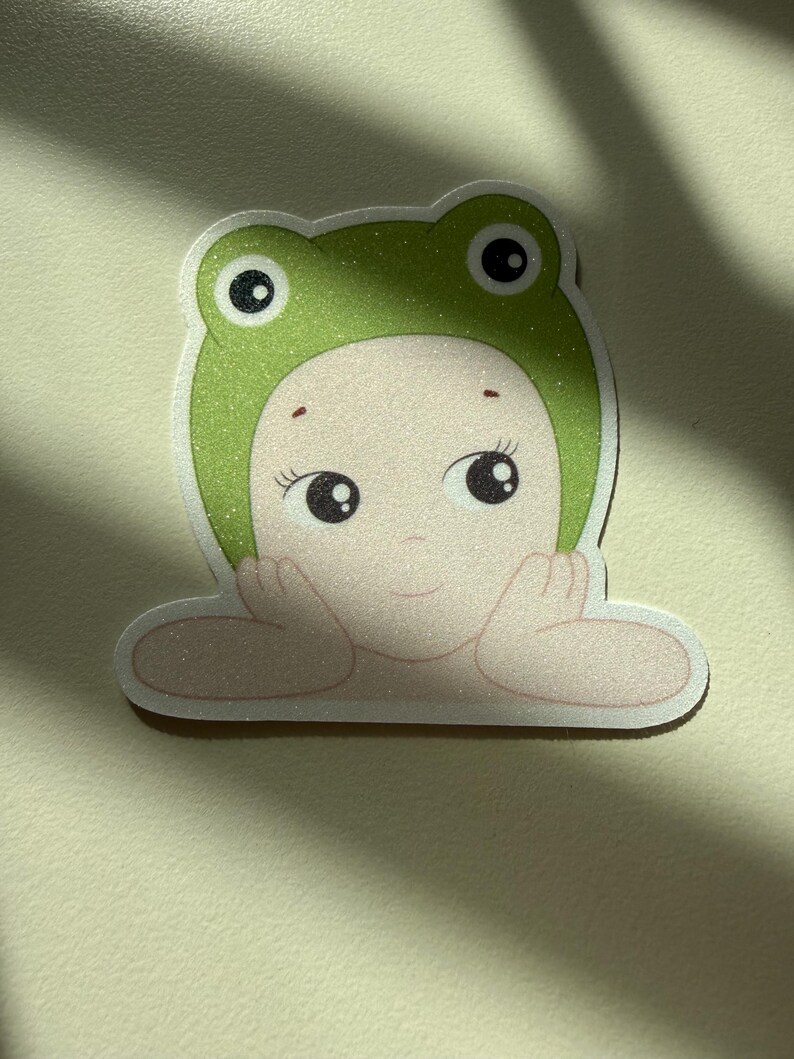 May include: A sticker featuring a cartoon illustration of a person wearing a green frog hat. The person has large, expressive eyes and is resting their chin on their hands. The hat has a glittery texture.