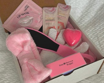 Puede incluir: Una caja de regalo de cuidado personal rosa abierta, llena de productos de belleza. Incluye una diadema rosa, una lima para pies, una esponja en forma de corazón, una mascarilla labial, sobres de exfoliante y una lima de uñas.