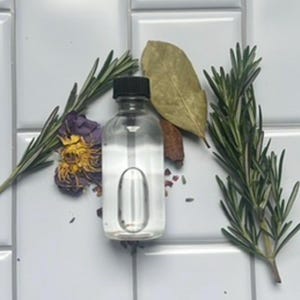 May include: A clear glass bottle with a black cap, surrounded by sprigs of rosemary, a bay leaf, a cinnamon stick, and dried flowers. The bottle has a unique oval-shaped indentation. The items are arranged on a white tiled surface.