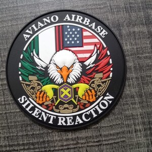 May include: A black and white patch with the text "Aviano Airbase Silent Reaction" around the edge. In the center is an eagle with its wings spread, holding an American flag and an Italian flag. The eagle is holding a purple and yellow symbol in its talons.
