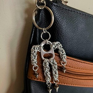 May include: A handmade silver-toned keychain featuring a chainmail figure with a bottle cap body. The keychain is attached to a black and brown leather bag with a silver clasp. The figure has three dangling cone-shaped accents.