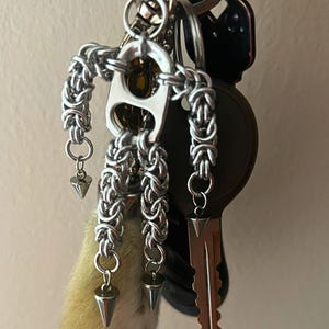 May include: A collection of keys on a silver keyring, including a car key and a house key. A handmade silver chain charm in the shape of a person is attached, along with three silver cone charms and a small, light-colored fur charm.