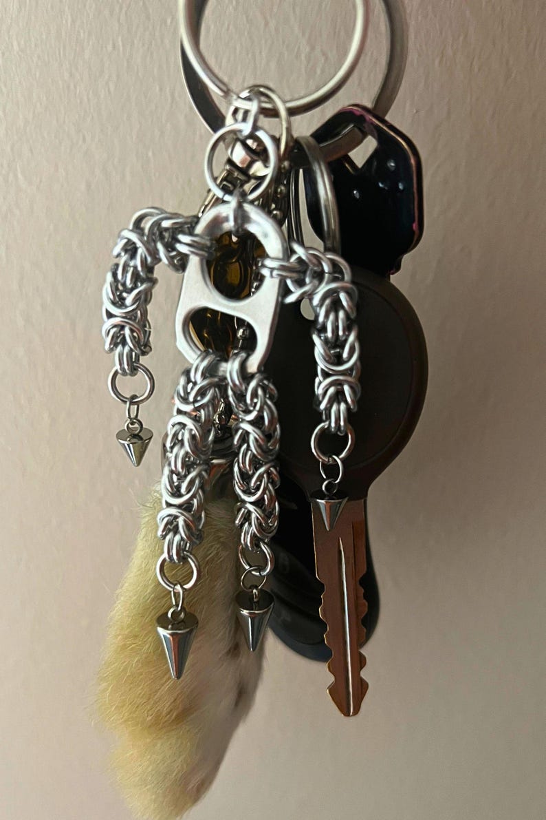 May include: A collection of keys on a silver keyring, including a car key and a house key. A handmade silver chain charm in the shape of a person is attached, along with three silver cone charms and a small, light-colored fur charm.