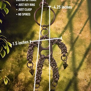 May include: A handmade, dark metal keychain featuring a stylized figure made of chainmail. The keychain is approximately 5.5 inches long, with a key ring and clasp. The figure has a 1.5-inch wide torso and 4.25-inch arms. Custom options include just a key ring, clasp, or no spikes.