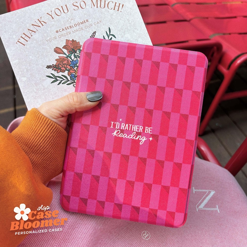 May include: A pink and white chequered e-reader case with the text "I'd rather be reading" in white. The case is held in a hand and the background is a pink surface.