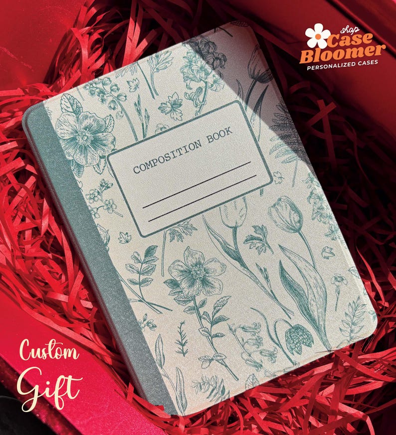 May include: A composition book with a teal and white floral pattern. The cover displays botanical illustrations of flowers and leaves. The words "COMPOSITION BOOK" are printed on a label. The book is presented in a red gift box with shredded paper.