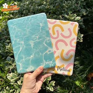 Könnte beinhalten: Zwei personalisierte Kindle E-Reader-Hüllen. Eine Hülle hat ein blaues und weißes wasserartiges Muster mit dem Namen "Ashley" darauf. Die andere Hülle hat einen weißen Hintergrund mit rosa und gelben abstrakten Formen und dem Namen "Melissa" darauf.