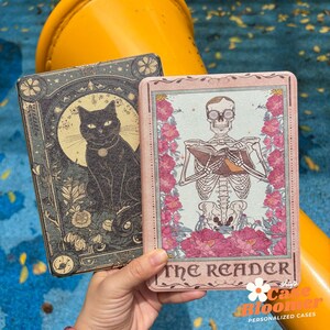 Könnte beinhalten: Zwei dekorative Handyhüllen. Eine zeigt eine schwarze Katze mit Mond- und Blumendesign. Die andere Hülle hat ein Skelett, das ein Buch liest, umgeben von rosa Blumen, mit dem Text "The Reader".