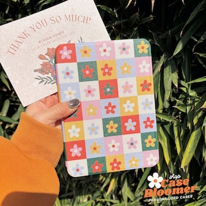 May include: A floral patterned case with a grid of colorful squares, each containing a flower. The case is held near a card that reads "THANK YOU SO MUCH!" and the shop name "Case Bloomer". The background is lush green foliage.