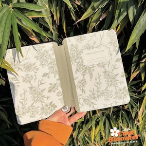 May include: Open composition book with a floral design on the cover. The cover is white with a green floral pattern. The book is open to a blank page with the words "Composition Book" written in a black frame.