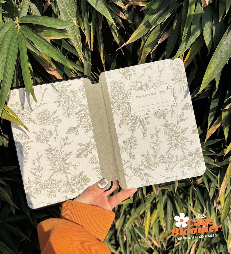 May include: Open composition book with a floral design on the cover. The cover is white with a green floral pattern. The book is open to a blank page with the words "Composition Book" written in a black frame.