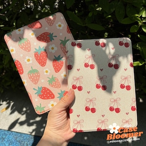 Könnte beinhalten: Zwei E-Reader-Hüllen mit unterschiedlichen Mustern. Eine Hülle hat einen rosa Hintergrund mit weißen Blüten und roten Erdbeeren. Die andere Hülle hat einen beigefarbenen Hintergrund mit roten Kirschen und weißen Schleifen. Beide Hüllen tragen den Text "Case Bloomer Personalized Cases".