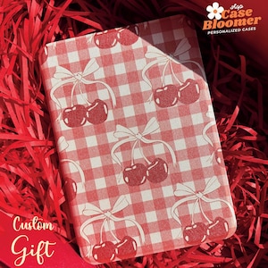 Puede incluir: Una funda de tel&eacute;fono a cuadros rojos y blancos con un dise&ntilde;o de cerezas y lazos. La funda est&aacute; en una caja de regalo roja con papel de seda rojo. El texto "shop Case Bloomer PERSONALIZED CASES" est&aacute; en la esquina superior derecha de la imagen. El texto "Custom Gift" est&aacute; en la esquina inferior izquierda de la imagen.