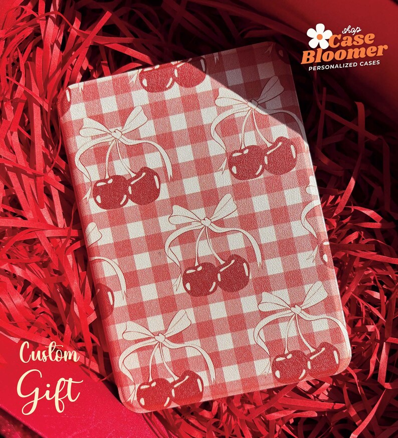 Puede incluir: Una funda de tel&eacute;fono a cuadros rojos y blancos con un dise&ntilde;o de cerezas y lazos. La funda est&aacute; en una caja de regalo roja con papel de seda rojo. El texto "shop Case Bloomer PERSONALIZED CASES" est&aacute; en la esquina superior derecha de la imagen. El texto "Custom Gift" est&aacute; en la esquina inferior izquierda de la imagen.