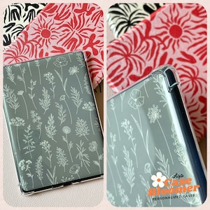 May include: A clear phone case with a gray floral design featuring white botanical illustrations. The case is shown with patterned fabric in the background, including red and black designs. The shop name "case bloomer" is visible.