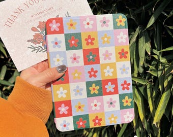 May include: A rectangular tablet case featuring a colorful checkerboard pattern of red, yellow, green, and light blue squares, each centered with a simple five-petaled flower. A white thank you card with floral illustrations is held behind the case.