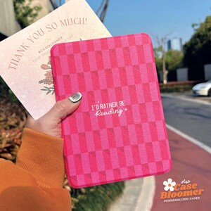 May include: A pink and white chequered e-reader case with the text "I'd rather be reading +" in white. The case is made of a soft, textured material and has a subtle sparkle.
