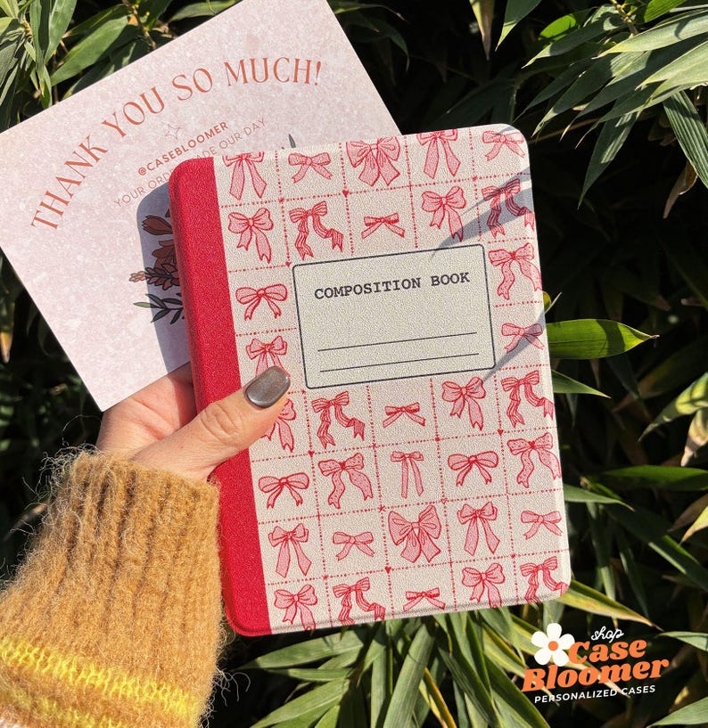 May include: A red and white composition book with a pink bow pattern. The book has the words "COMPOSITION BOOK" printed on the front cover.