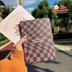 May include: A brown and tan chequered case with the text "my weekend is BOOKED" in gold lettering. A thank you card with floral illustrations is held next to the case. The shop name "Case Bloomer" is visible.
