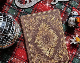 May include: A brown leather-bound journal with gold ornate designs and a crescent moon symbol on the cover. The journal is on a red and green plaid fabric with a silver glitter "Merry Christmas" sign and a snowman figurine.