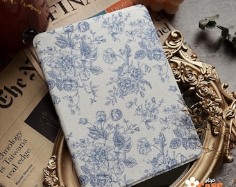 May include: A white and blue floral patterned tablet case with a white background and blue floral designs. The case is made of a soft, textured material. The case is shown on a table with a gold frame and a magazine.