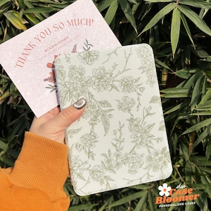 May include: A light green notebook cover with a floral pattern. The cover has a white background with green floral illustrations. The text "shop Case Bloomer PERSONALIZED CASES" is printed on the bottom of the cover.