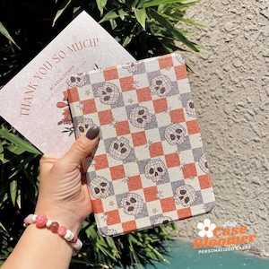 May include: A patterned case featuring a checkerboard design with skull graphics. The case is held with a card that reads "THANK YOU SO MUCH!" and a floral design. The shop name "Case Bloomer" is visible in the bottom right corner.