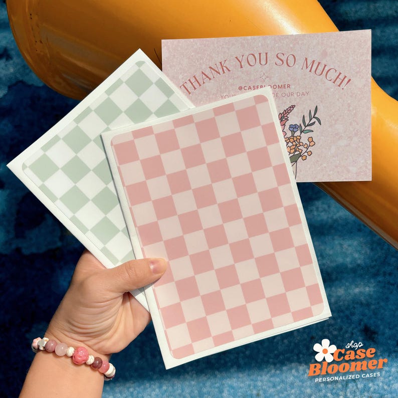 May include: A hand holds two checkered pattern cases, one pink and one light green, with a thank you card. The card has the text "THANK YOU SO MUCH!" and a floral design. The shop logo "Case Bloomer" is in the bottom right.