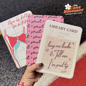 Op de afbeelding: Drie gepersonaliseerde telefoonhoesjes met unieke ontwerpen. Eén hoesje heeft een bh op een waslijn met de tekst "life is better in a bikini". Een andere heeft een roze patroon met het woord "smut". De derde is ontworpen als een bibliotheekkaart met de tekst "buy me books & tell me I'm pretty."