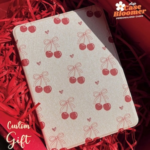 May include: A white custom case with a red cherry pattern and a bow on each cherry. The case has the text "shop Case Bloomer PERSONALIZED CASES" in the top right corner. The case is sitting on a red shredded paper background.