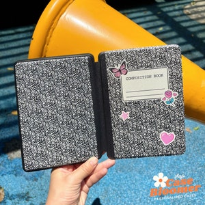 Op de afbeelding: Een zwart-witte e-reader hoes in de stijl van een schrift met een roze vlinder, ster en hart stickers. De hoes heeft de tekst "Composition Book" op de voorkant.