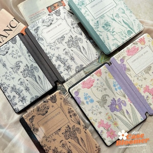 May include: Several composition book covers with floral designs. The covers display botanical illustrations of irises, roses, and other flowers. Some covers have the words "COMPOSITION BOOK" printed on them. The colour palette includes shades of beige, green, and purple.