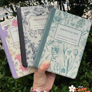 May include: Three composition books with floral patterns. The covers display diverse colours and designs, including pink, blue, and black. Each book has a label that reads "COMPOSITION BOOK". The books are held against a green leafy backdrop.