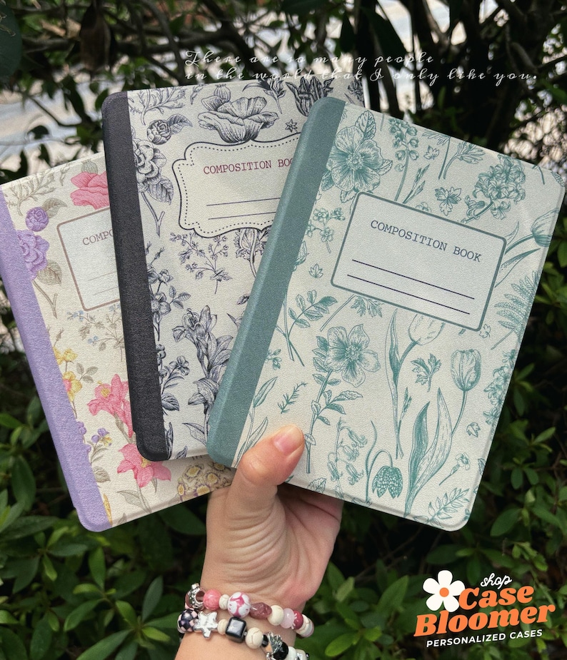 May include: Three composition books with floral patterns. The covers display diverse colours and designs, including pink, blue, and black. Each book has a label that reads "COMPOSITION BOOK". The books are held against a green leafy backdrop.